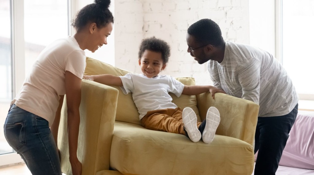 Parents moving an armchair with a little boy sitting on it. 