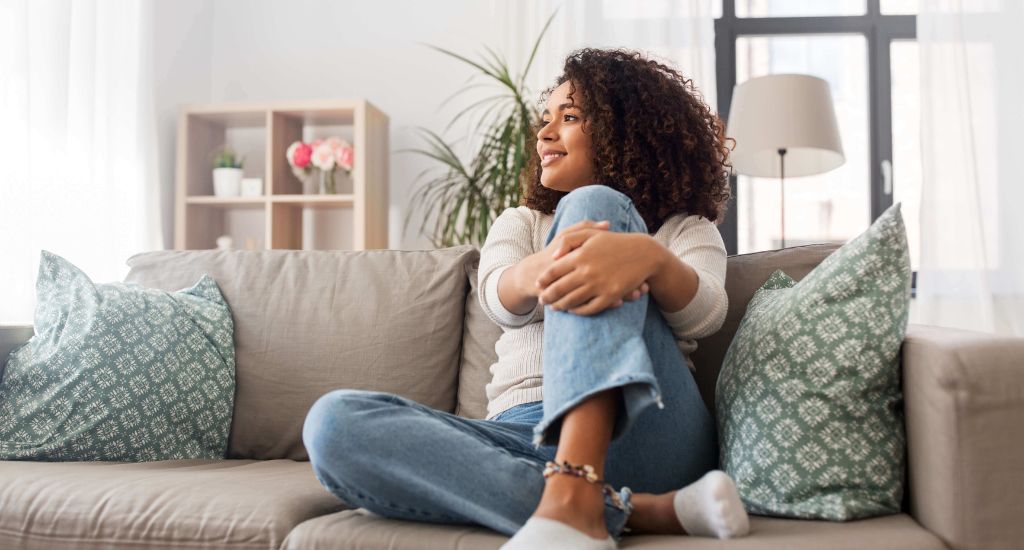 Femme assise sur un sofa dans un salon lumineux qui regarde sereinement par la fenêtre