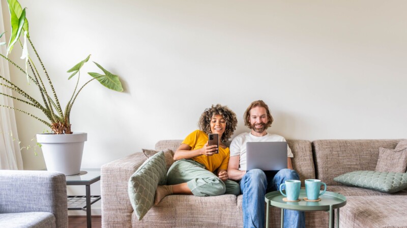 Couple assis sur un sofa, regardant leurs écrans en souriant - Taux fixe ou taux variable : comment choisir le bon taux hypothécaire? | Multi-Prêts Hypothèque